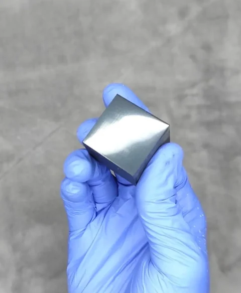 A dark cube held in a blue glove, top face polished to a mirror reflection